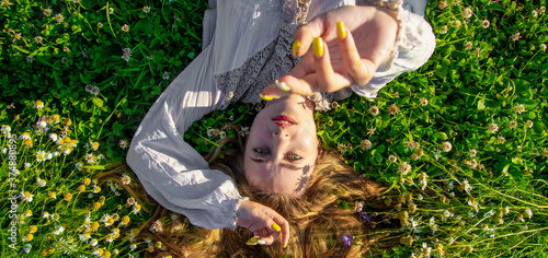 a young girl lies on the grass and wildflowers on her back and reaches out to the frame forward. focusing on the face. photo top view.