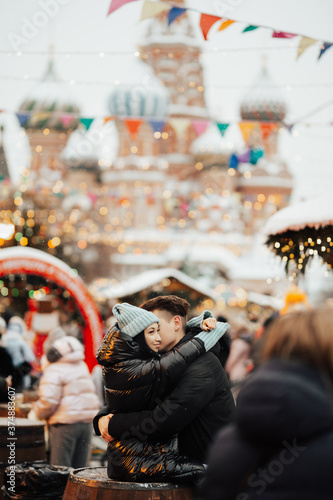 Young romantic couple in warm clothes hugs on the street at the festival on Christmas day, enjoying spending time together.