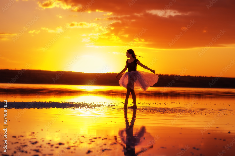 Girl ballerina in a ballet dress dances in the water of the lake at sunset. Silhouette of a slender girl in a transparent airy dress against the backdrop of the setting sun.