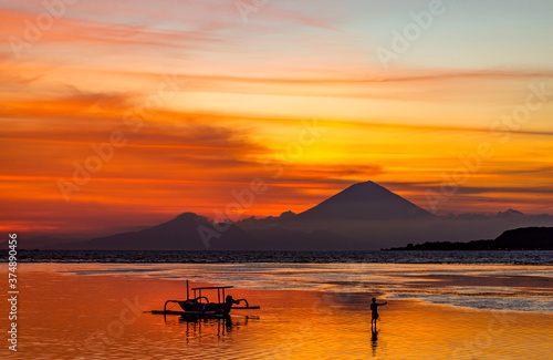 Silhouette of a person and a small traditional boat against sunset over the sea with a volcano in the distance