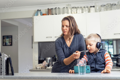Woman in the kitchen feeds her little son and watching at electronic tablet. Child sits on table in headphones and watch at mobile phone