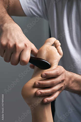 Male masseur hands doing massage on female foot reflex zone in the spa salon using gua sha scraping acupuncture tool.