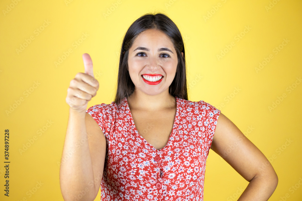 Young beautiful woman over isolated yellow background smiling and doing the ok signal with her thumb
