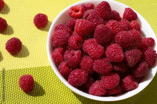 Raspberries in a white bowl on a green background. Ripe berries are scattered.