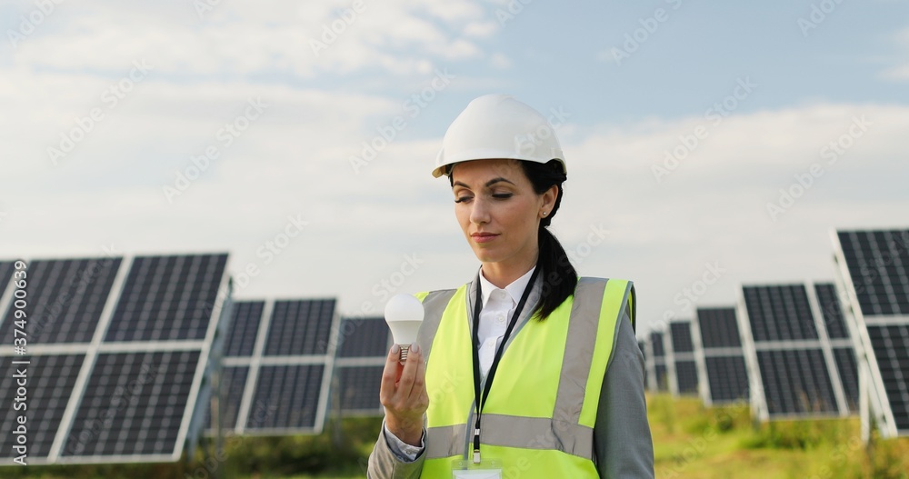Portrait of electrician engineer in safety helmet and uniform near photovoltaic solar panels. Female engineer holding bulb on solar panel background.
