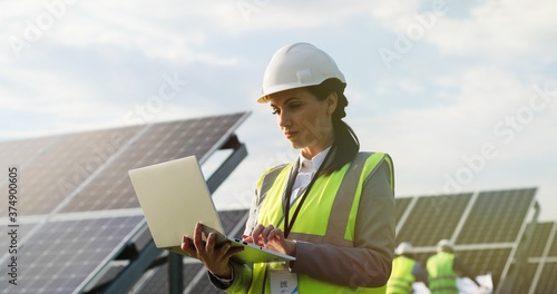 Portrait of electrician engineer in safety helmet and uniform using laptop checking solar panels. Female technician at solar station. photo