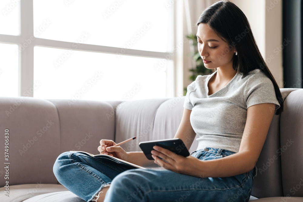 Image of young asian woman using mobile phone while writing down notes Image of young asian woman using mobile phone while writing down notes