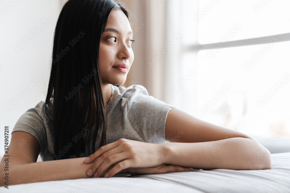 Image of pleased nice asian woman looking aside while sitting on sofa Image of pleased nice asian woman looking aside while sitting on sofa