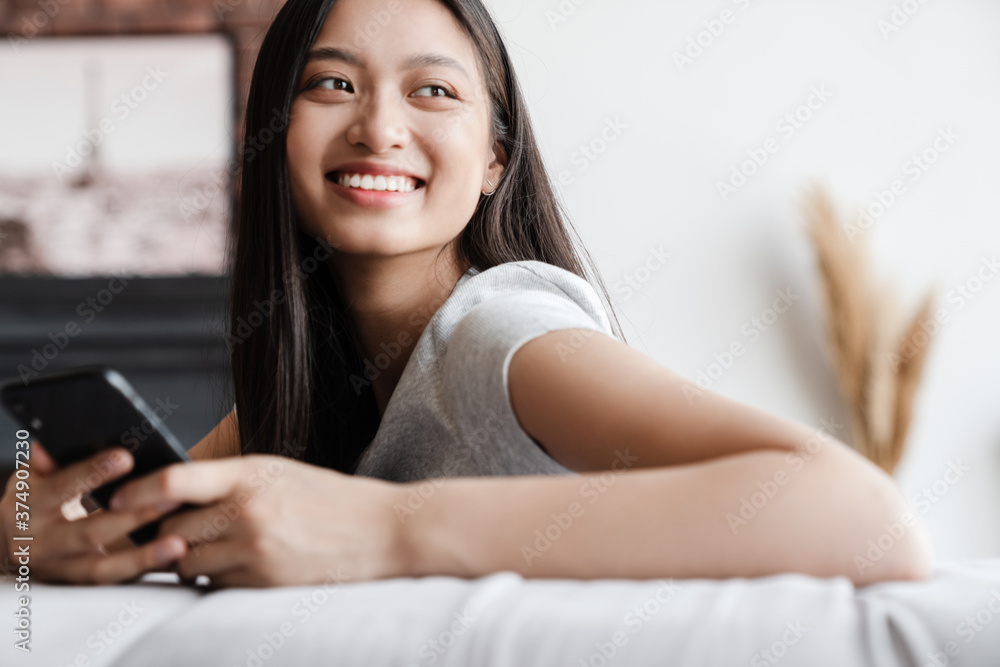 Image of happy asian woman using mobile phone while sitting on sofa Image of happy asian woman using mobile phone while sitting on sofa