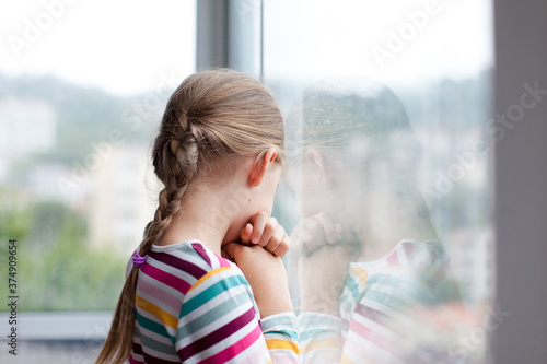 Sad child looking through rainy window at home. Upset kid in self isolation during quarantine. Lifestyle, authentic, candid moment. photo