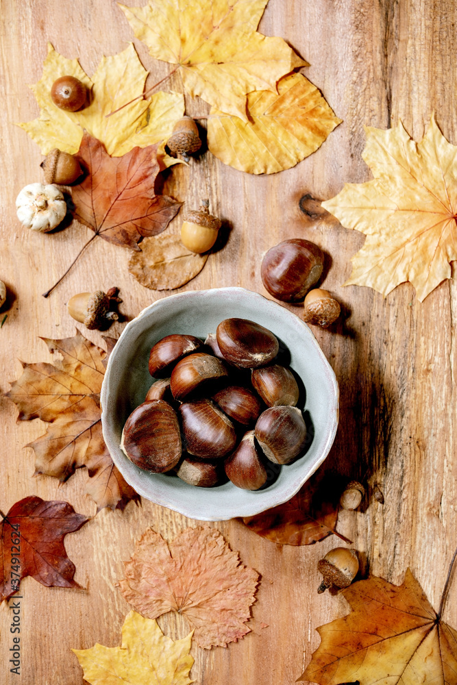 Raw edible chestnuts in ceramic bowl and yellow autumn maple leaves over wooden texture background. Flat lay