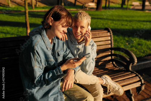Two joyful girls using smartphone, sitting on the bench in the city park. Lesbian couple holding their phones while spending time together outdoors