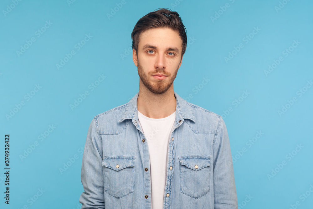 Portrait of serious handsome young man with stylish hairdo and bristle, wearing worker denim shirt looking at camera with calm attentive focused expression. studio shot isolated on blue background Portrait of serious handsome young man with stylish hairdo and bristle, wearing worker denim shirt looking at camera with calm attentive focused expression. studio shot isolated on blue background