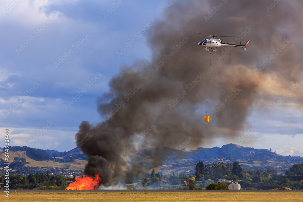 Fototapeta premium A helicopter with a water bucket going to put out a fire by an airport runway