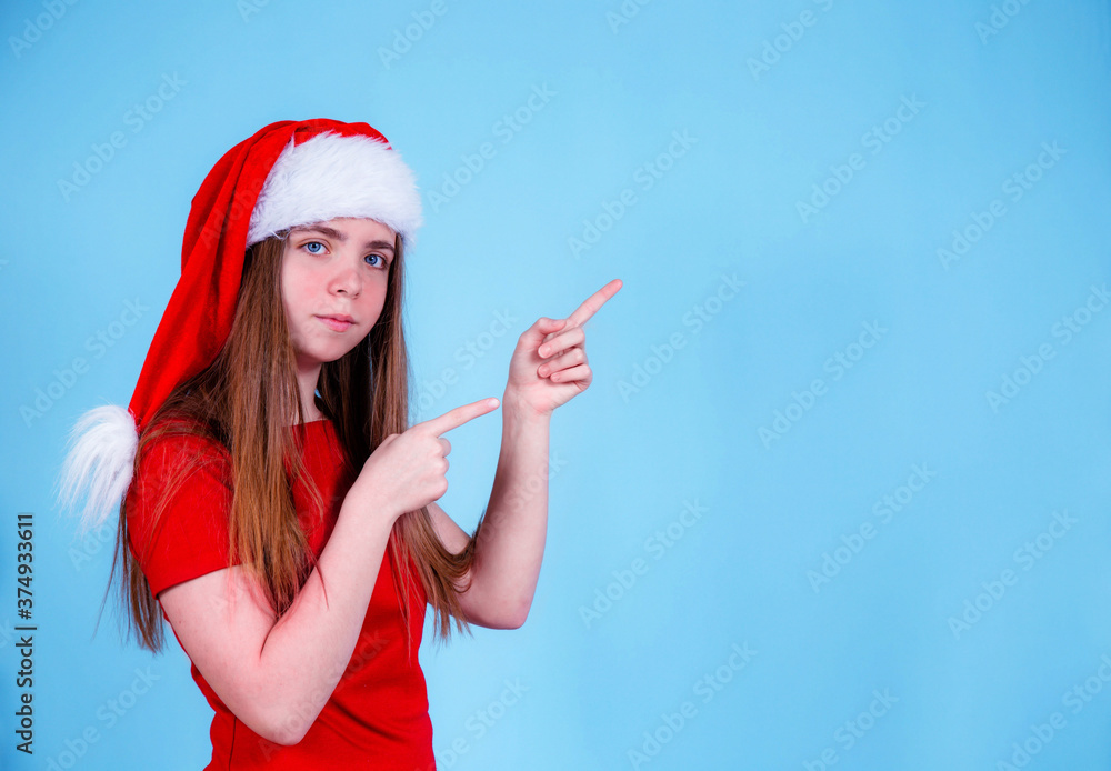 Fototapeta premium a young girl in a Santa Claus hat on a blue background shows where to look.