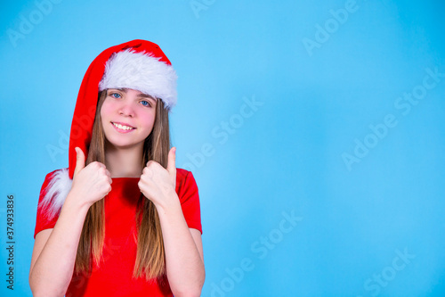 Beautiful girl in a Christmas Santa hat on a blue background with a happy smiling face makes