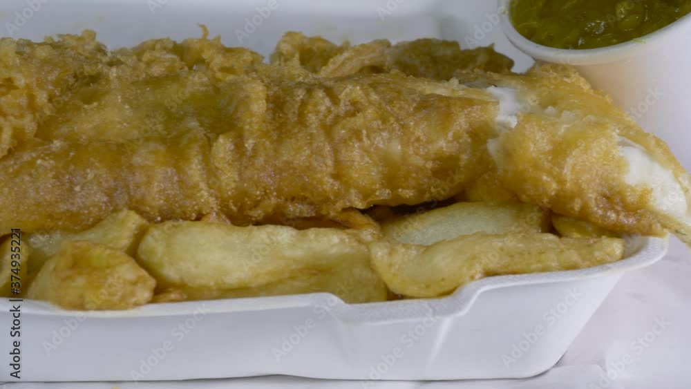 Dolly shot of traditional British takeaway hot meal of deep fried, white fish and chips and batter, in a carton and paper wrapping, with a pot of thick curry sauce, opened up ready to eat. Dolly shot of traditional British takeaway hot meal of deep fried, white fish and chips and batter, in a carton and paper wrapping, with a pot of thick curry sauce, opened up ready to eat.