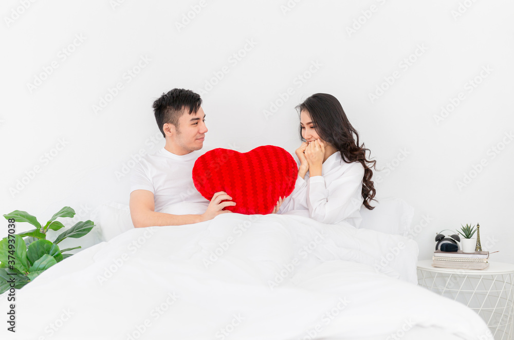 asian man holding red heart pillow and send to asian female, they rest on white bed, happy valentines day asian man holding red heart pillow and send to asian female, they rest on white bed, happy valentines day