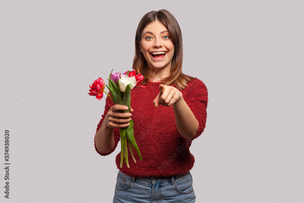 Flowers for you! Portrait of cheerful happy brunette girl smiling, holding tulips bouquet and pointing at camera. Congratulations on spring holidays. indoor studio shot isolated on gray background Flowers for you! Portrait of cheerful happy brunette girl smiling, holding tulips bouquet and pointing at camera. Congratulations on spring holidays. indoor studio shot isolated on gray background