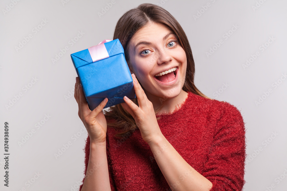 Excited happy girl in shaggy sweater holding gift box near ear, listening guess what's inside, in anticipation of christmas present, birthday surprise. indoor studio shot isolated on gray background Excited happy girl in shaggy sweater holding gift box near ear, listening guess what's inside, in anticipation of christmas present, birthday surprise. indoor studio shot isolated on gray background