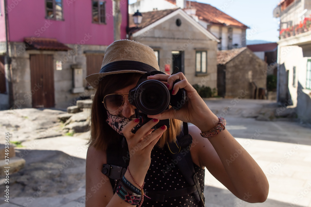 Attractive young female taking a photo with a reflex camera while wearing a hat and a face mask and visiting an old town in Extremadura, Spain
