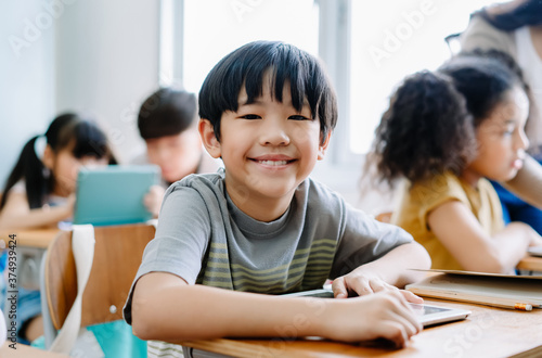 Cute little Asian boy smiling looking at camera and using laptop in computer class at the elementary school. Education, school, technology and internet concept.