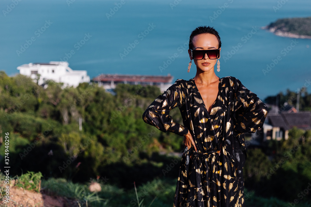 Fototapeta premium Portrait of beautiful luxury look fit tanned slim woman in black golden cape and sunglasses on top of mountain with amazing sea view in Thailand. 