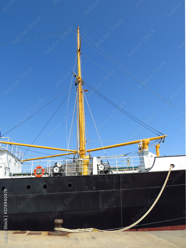 Side view of merchant ship hull and its rigging moored at industrial dock under intense blue sky. Maritime navigation and transport.