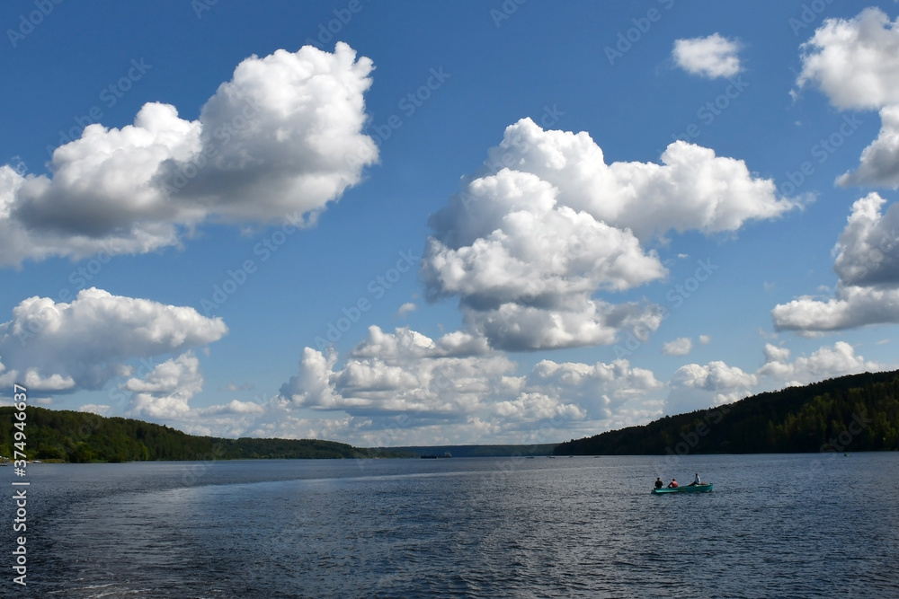 Blue sky with beautiful white clouds over a wide river. Ripples on the surface of the water. Low cloud. Green forest on the banks. A small boat in the middle of the river.