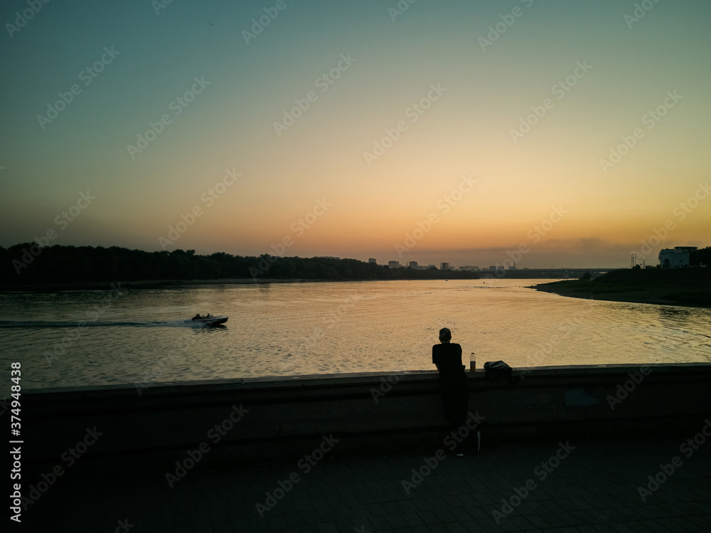 A man is standing on the embankment and watching a boat passing by in the sunset