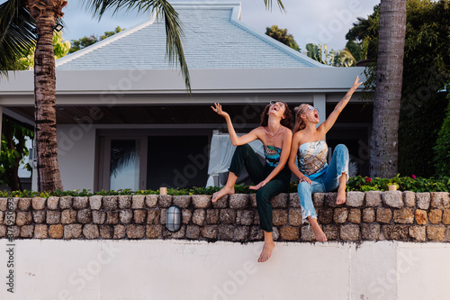 Two happy woman friends with sunglasses on vacation in tropical country. Females in top and jeans having fun at villa hotel, palm trees on background. 