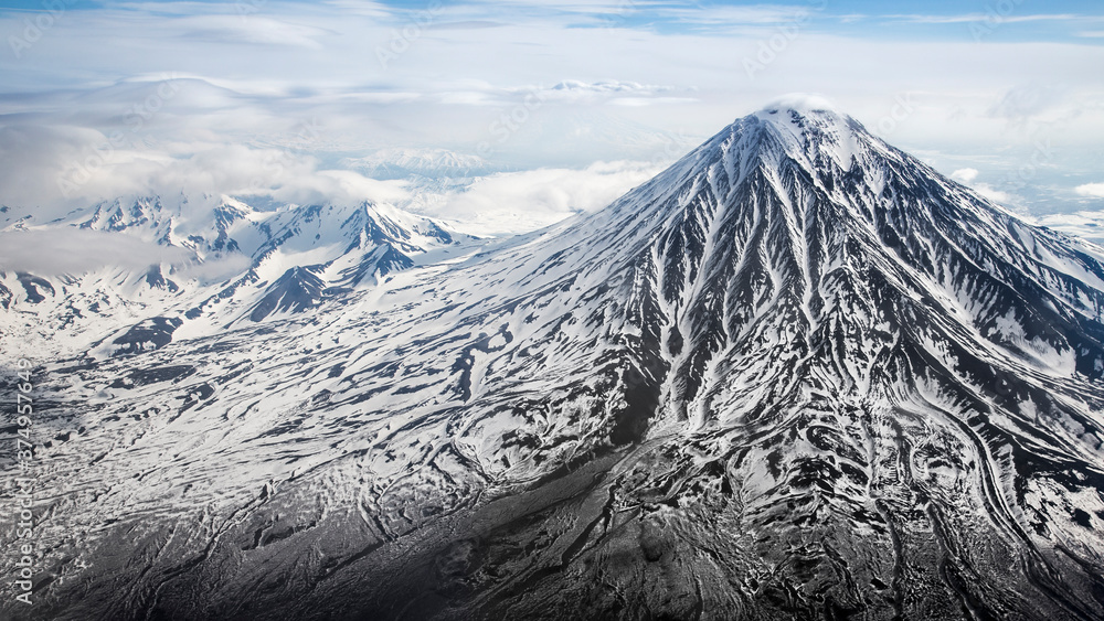 A black volcano with streaks of white snow on a background of mountain peaks and clouds. Kamchatka, a shot from an airplane.
