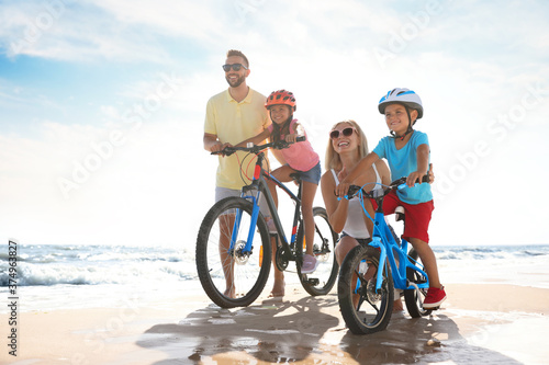 Happy parents teaching children to ride bicycles on sandy beach near sea