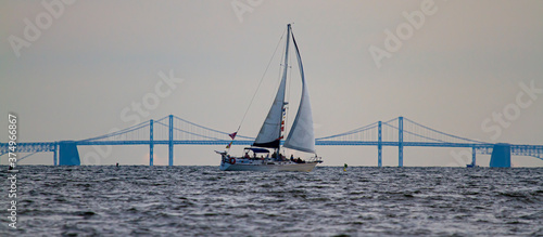 Panoramic image of a sailboat moving across Chesapeake Bay with the silhouette of the famous Bay Bridge in the background. There are people on the boat who are enjoying the sunset over the bay,.