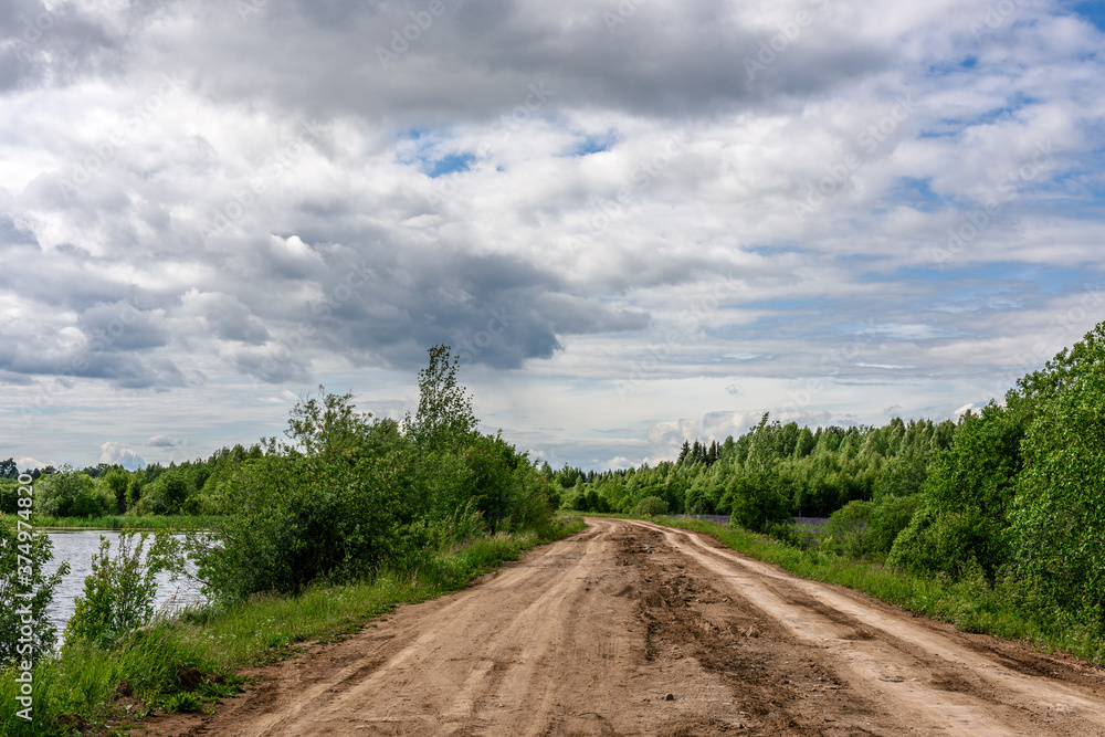 Dirt road against blue sky, green forest and lake.