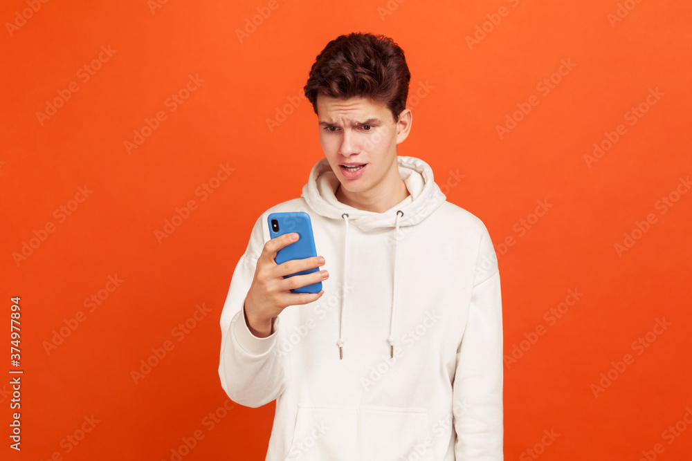 Wondered teenager in white casual sweatshirt with hood looking at smartphone screen with amazed face, bad connection. Indoor studio shot isolated on orange background Wondered teenager in white casual sweatshirt with hood looking at smartphone screen with amazed face, bad connection. Indoor studio shot isolated on orange background