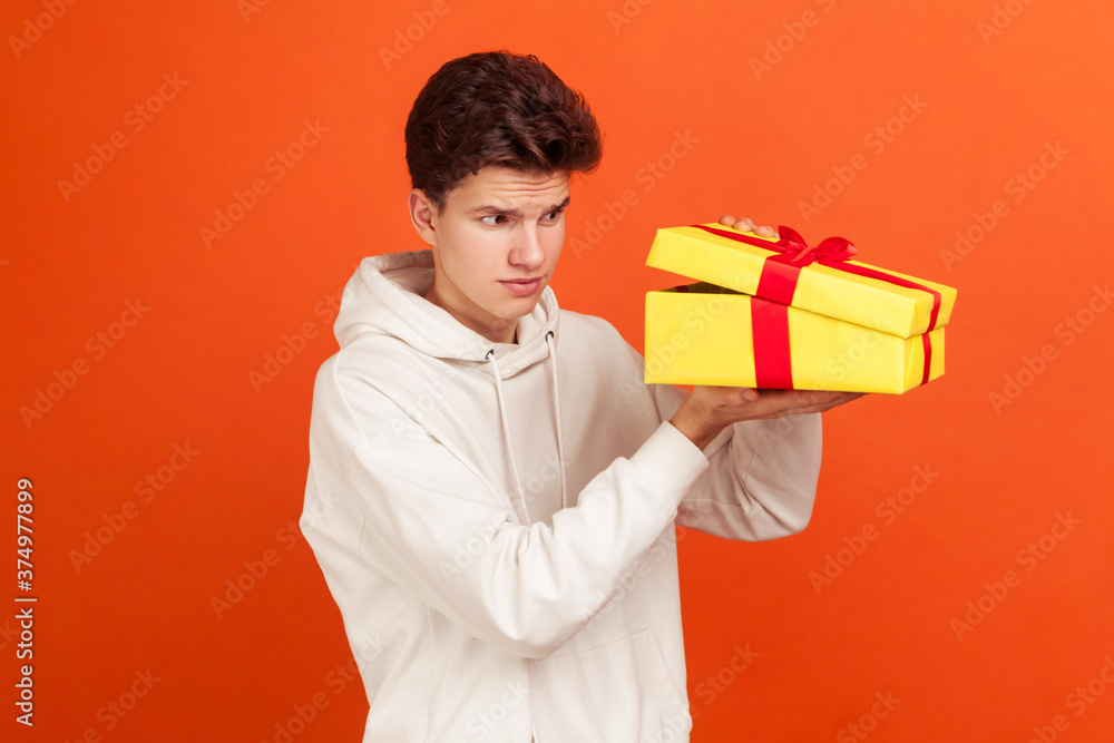Profile portrait of doubtful teenager with fashionable hairdo in casual sweater looking inside gift box, peeping. Indoor studio shot isolated on orange background Profile portrait of doubtful teenager with fashionable hairdo in casual sweater looking inside gift box, peeping. Indoor studio shot isolated on orange background