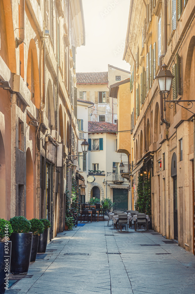 Fototapeta premium Typical italian street with old colorful buildings with shutter windows, street lights at wall, restaurant tables, Brescia city historical centre, vertical view, Lombardy, Northern Italy