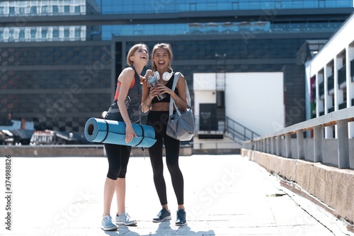 Two happy smiling young athletic woman walking outdoors together after sport training, talking and hugging each other