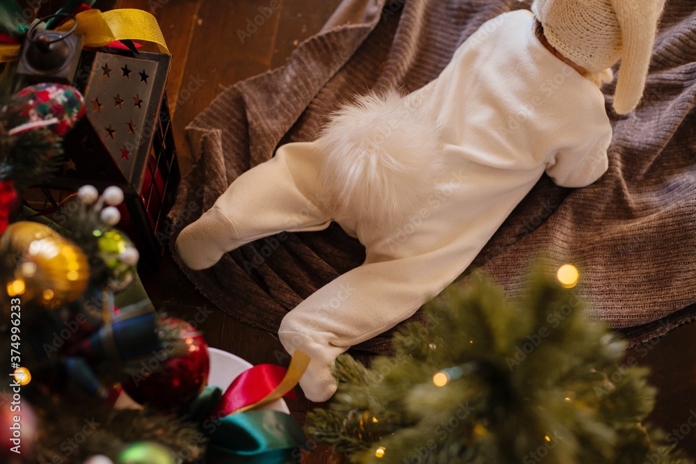 A newborn baby lies on a blanket near the Christmas tree in a rabbit costume tail up, face down. A kid in a rabbit costume crawls on the veil.