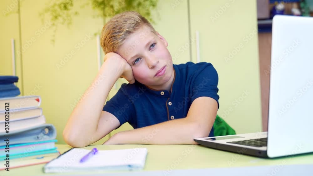 Cute tired schoolboy with sad face sitting at the table with notebooks and laptop during online studying isolated in at home. Boring teenager hold his head, during e-learning, wearing blue t-shirt. Cute tired schoolboy with sad face sitting at the table with notebooks and laptop during online studying isolated in at home. Boring teenager hold his head, during e-learning, wearing blue t-shirt.