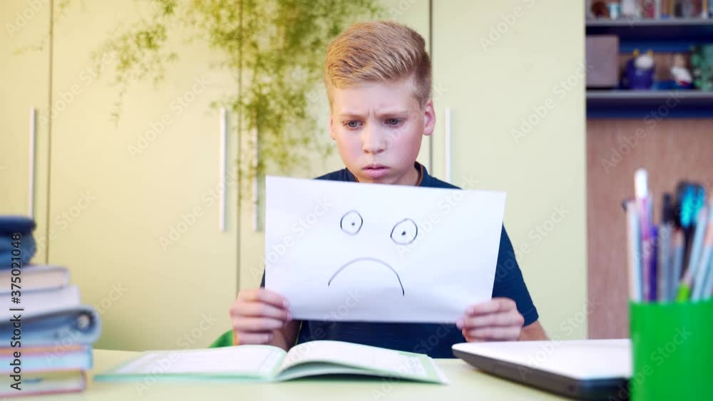 portrait of a dissatisfied young schoolboy boy opens a notebook and shows a smile unhappy and sad. A teenager at a table in a classroom or room reads a book and looks at a task in a notebook. portrait of a dissatisfied young schoolboy boy opens a notebook and shows a smile unhappy and sad. A teenager at a table in a classroom or room reads a book and looks at a task in a notebook.