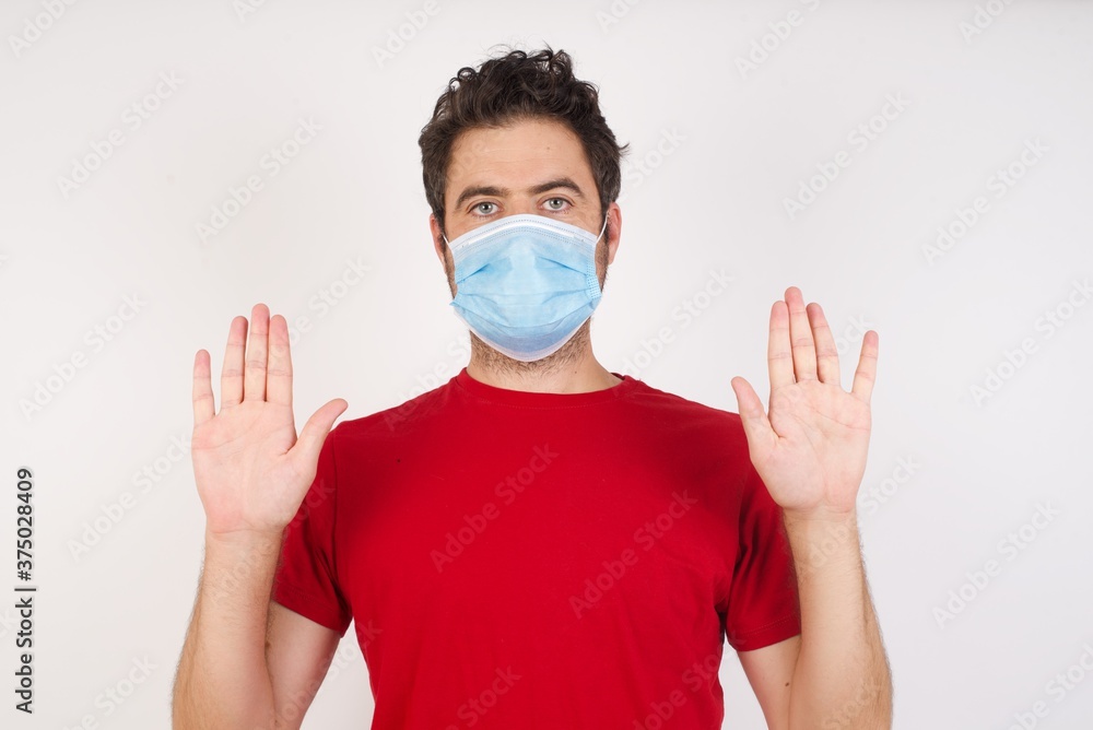 Serious Young caucasian man with short hair wearing medical mask standing over isolated white wall pulls palms towards camera, makes stop gesture, asks to control your emotions and not be nervous Serious Young caucasian man with short hair wearing medical mask standing over isolated white wall pulls palms towards camera, makes stop gesture, asks to control your emotions and not be nervous