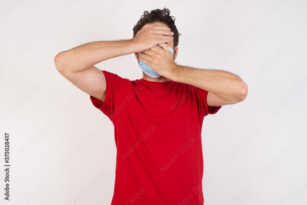 Young caucasian man with short hair wearing medical mask standing over isolated white background Covering eyes and mouth with hands, surprised and shocked. Hiding emotions. Young caucasian man with short hair wearing medical mask standing over isolated white background Covering eyes and mouth with hands, surprised and shocked. Hiding emotions.