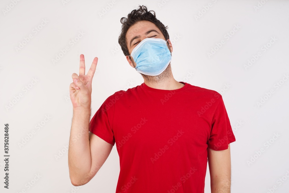 Young caucasian man with short hair wearing medical mask standing over isolated white background smiling with happy face winking at the camera doing victory sign. Number two. Young caucasian man with short hair wearing medical mask standing over isolated white background smiling with happy face winking at the camera doing victory sign. Number two.