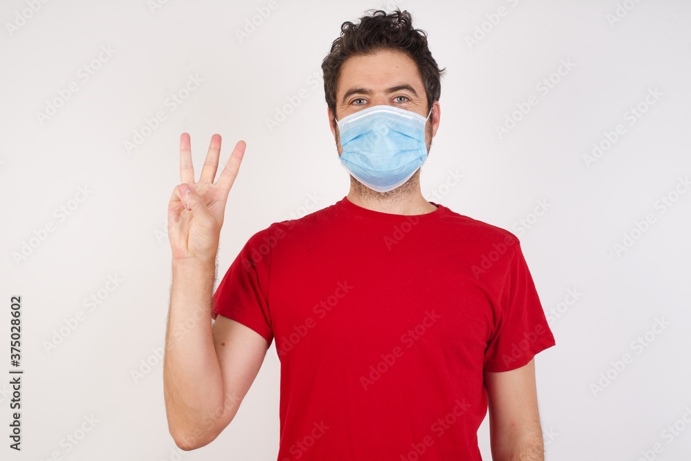 Young caucasian man with short hair wearing medical mask standing over isolated white background showing and pointing up with fingers number three while smiling confident and happy. Young caucasian man with short hair wearing medical mask standing over isolated white background showing and pointing up with fingers number three while smiling confident and happy.