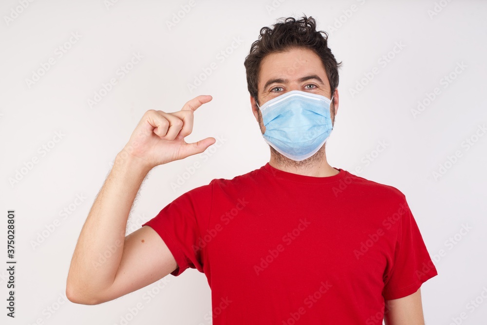 Young caucasian man with short hair wearing medical mask standing over white wall smiling and confident gesturing with hand doing small size sign with fingers looking and the camera. Measure concept