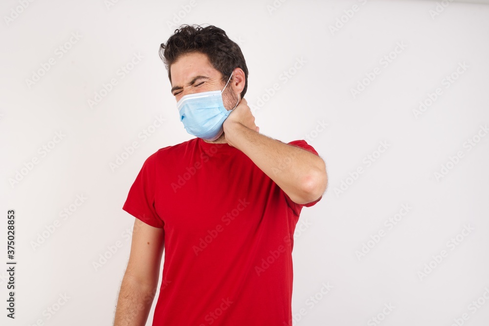 Young caucasian man with short hair wearing medical mask standing over isolated white background Suffering of neck ache injury, touching neck with hand, muscular pain Young caucasian man with short hair wearing medical mask standing over isolated white background Suffering of neck ache injury, touching neck with hand, muscular pain