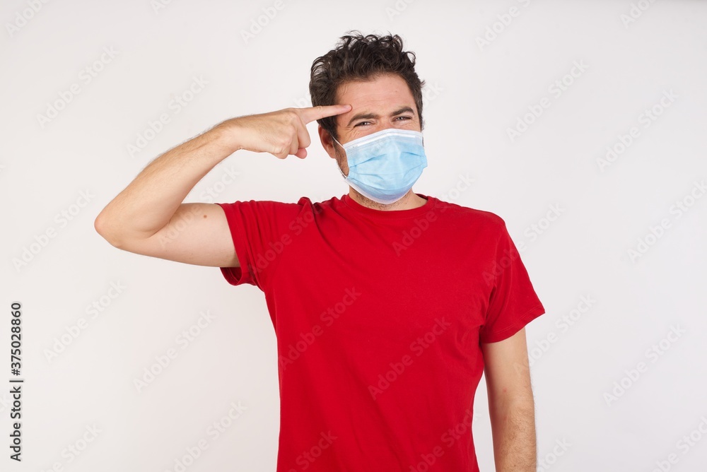 Young caucasian man with short hair wearing medical mask standing over isolated white background pointing unhappy at pimple on forehead, blackhead infection. Skincare concept. Young caucasian man with short hair wearing medical mask standing over isolated white background pointing unhappy at pimple on forehead, blackhead infection. Skincare concept.