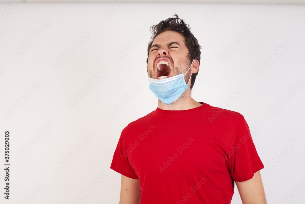 Young caucasian man with short hair wearing medical mask standing over isolated white background angry and mad screaming frustrated and furious, shouting with anger. Rage and aggressive concept.